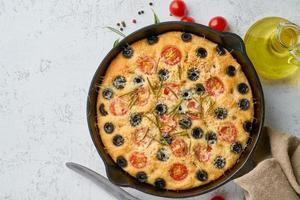 Focaccia, pizza in skillet, italian flat bread with tomatoes, olives and rosemary. Top view, copy space, white concrete background photo