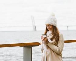 Beautiful young girl drinking coffee, tea from plastic mug in autumn, winter. A woman with long hair stands on waterfront on Baltic sea in port and waiting for ferry, heated by hot drink, copy space photo