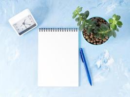 Top view of modern bright blue office desktop with notepad, clock, flower succulent. Mock up, empty space photo