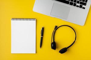 Modern office desktop. Blank notepad, pen, headset and laptop on yellow background