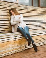 Beautiful young girl with long brown hair sits on wooden bench made of planks and rests, sleeps or dozes in fresh air. Outdoor photo shoot with attractive woman in winter or autumn.