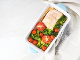 Fish salmon baked in oven with vegetables - broccoli, tomatoes. Healthy diet food, white marble backdrop, top view. photo