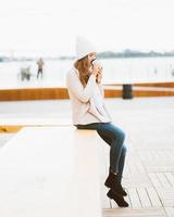 Beautiful young girl drinking coffee, tea from plastic mug in autumn, winter. A woman with long hair sitting on waterfront on Baltic sea in port and waiting for ferry, heated by hot drink, vertical