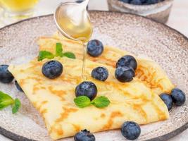 Two pancakes with blueberries and honey on plate, honey flows from spoon, side view, close-up photo