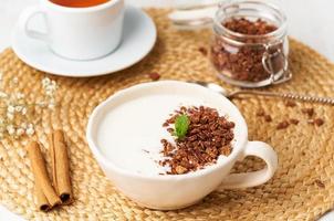 Yogurt with chocolate granola in cup, breakfast with tea on beige background, side view. photo