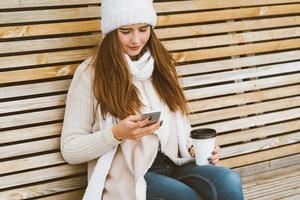Beautiful young girl drinking coffee, tea from plastic mug in autumn, winter and talking on mobile phone. Woman with long hair sitting on bench in autumn or winter, basking in hot drink, copy space photo