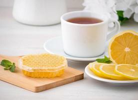 taza blanca con té, rodajas de limón y miel en panal sobre mesa de madera blanca, vista lateral foto