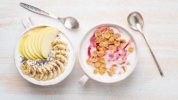 dos yogures griegos con mermelada, manzana, muesli, semillas de chía y plátano en un tazón blanco sobre una mesa de madera blanca, vista superior, pancarta foto