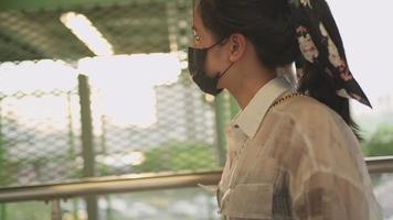 jonge aziatische vrouw met een zwart masker die alleen loopt op het voetgangerspad van de brug met zonsondergang op de achtergrond, nieuw normaal leven als je naar buiten gaat, zelfpreventie, bescherm jezelf tegen covid-19 corona-virus