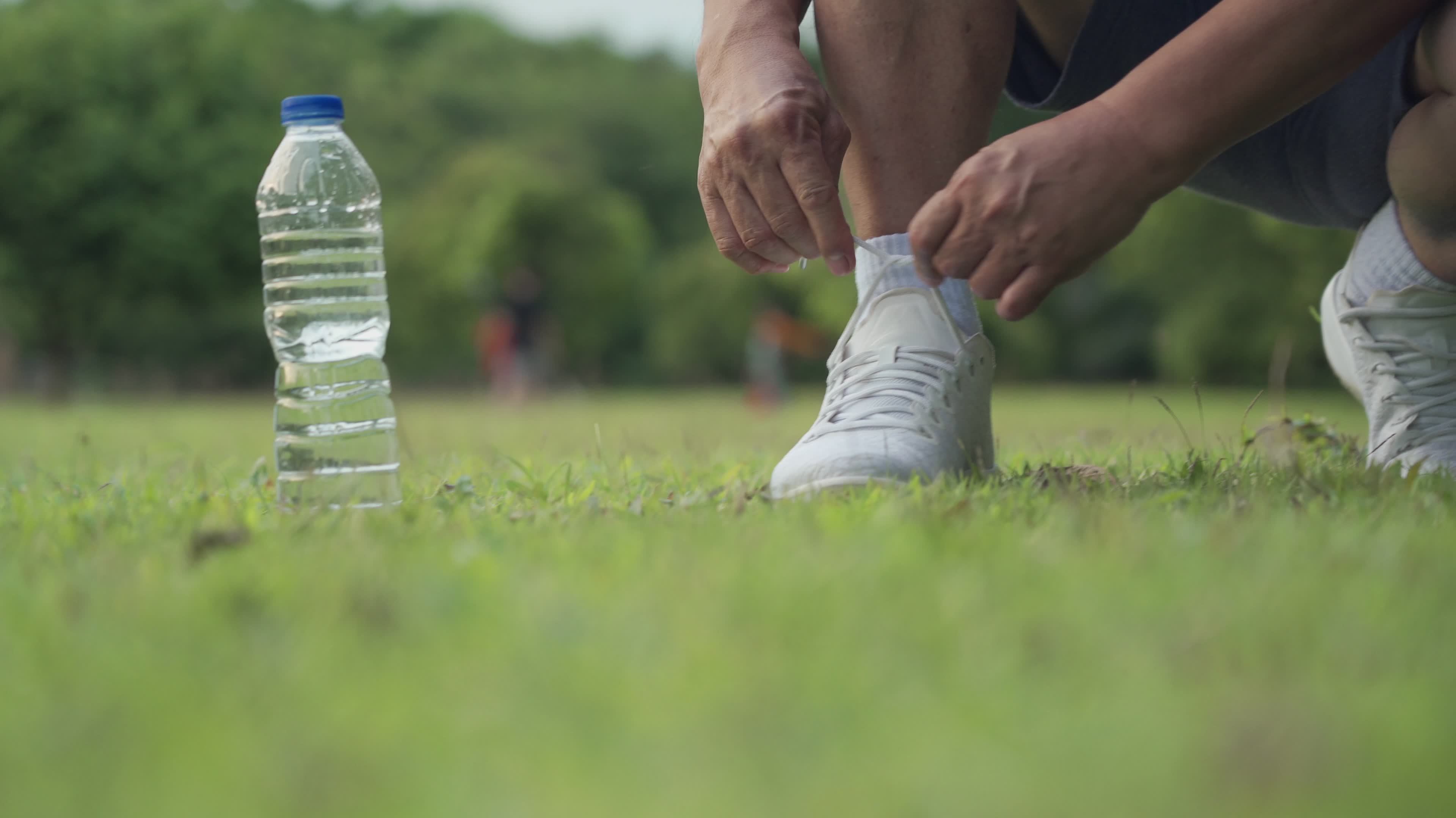 Asian male sitting down tie shoes laces on green grass field during  Exercise at the park with trees Background, outdoor exercising, active  lifestyle, stay hydrated water bottle on the side, front view