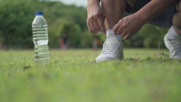 Asian male sitting down tie shoes laces on green grass field during Exercise at the park with trees Background, outdoor exercising, active lifestyle, stay hydrated water bottle on the side, front view video