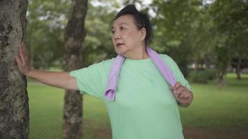 donna anziana asiatica che corre e si esercita al parco, in piedi accanto all'albero e riposa, benessere del corpo umano, assistenza sanitaria femminile, piano di assicurazione sulla vita, routine di allenamento, buona salute per una vita migliore video