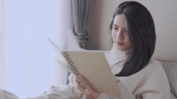 jovem mulher asiática sente-se na cama lendo livro sozinha, tarefa matinal produtiva, passando o tempo livre no fim de semana, luz do dia da janela do quarto confortável, auto-educação, histórias de literatura de autor video