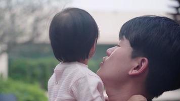 Head shot of young Asian father holding and kissing his adorable cute baby girl, small kid child care parenting, standing outside shot, Family member bonding, daddy lift baby up rest on his shoulder video