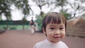 asiatiskt ungt kvinnligt barn som går själv i offentlig park, pappa väntar i bakgrunden, småbarns första steg, början på nytt liv, söt och energisk liten flicka i parken under träd video