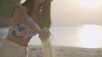 jovem mulher asiática brincando com areia na praia, pegando areia branca e mostrá-la para a câmera, derramando areia garota despreocupada com chapéu de batida, sente-se na ilha de praia tropical, reflexo do sol no oceano video