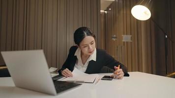 Focused asian secretary woman works on smartphone app, laptop computer on working desk alone in night time. Female professional writes down an important information on paperwork, distant communication video