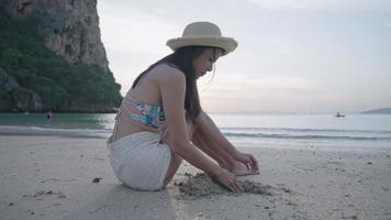 draaien close-up shot een jonge speelse aziatische vrouw zit knieën omhoog aan het strand, handen graven in een schoon zand, een meisje in sexy strandkleding plezier spelend alleen onder mensen, vrouw op solo reisconcept