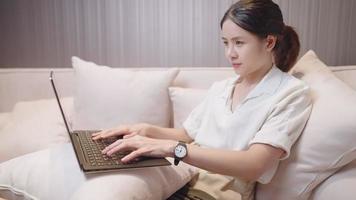 mulher asiática feliz usando laptop dentro de uma sala de estar confortável, trabalhar em casa, colocando o laptop no travesseiro, hábito de compras on-line, local de trabalho remoto do apartamento no sofá aconchegante, tecnologia sem fio