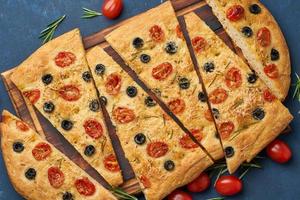 Focaccia, pizza, italian flat bread with tomatoes, olives and rosemary on dark blue table, top view