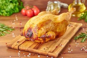 Whole roasted chicken on wooden cutting board on dark brown wooden table, side view photo