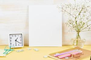 Mockup with blank white frame on yellow table against wooden wall, alarm, flower in vaze photo