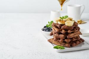 Chocolate banana waffles with maple syrup on white table, copy space, side view. Sweet brunch, maple syrup drop. photo