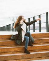 Beautiful young woman in warm clother is talking on phone and pointing with her hand. Girl sits on wooden steps on waterfront in port in autumn, winter, vertical