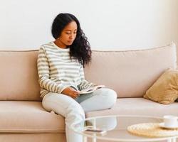 Beautiful African-American female sitting on couch and reading book. Spending time at home alone