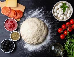 Raw dough for pizza with ingredients and spices on dark black background, top view, flat lay photo