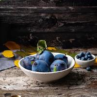 autumn background with ripe violet blue plums in white bowl on old grey wooden table, side view photo