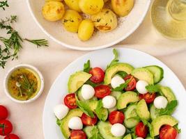 Fresh salad. Tomatoes, cucumbers, arugula, mozzarella, avocado. Fragrant oil with rosemary thyme, baked potato, top view