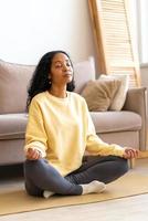 Young African-American female sitting in lotus yoga pose on mat in while meditating, vertical photo