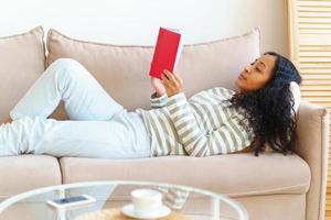 African-American lying down on sofa and reading book in red cover. Slow paced lifestyle photo