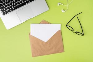 Concept of open craft envelope with blank paper sheet on green background. Laptop, earphones photo