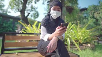 Asian Muslim female wear hijab and protective face mask. using smartphone sitting down relaxing time inside public park, New normal COVID pandemic, outdoors sunny day, wireless technology connection video