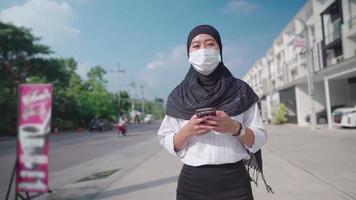 jovem mulher asiática em hijab e máscara facial protetora usando mensagem de texto para smartphone, andando na beira da estrada com conexão de rede de bairro urbano, pandemia covid-19 novo distanciamento normal video