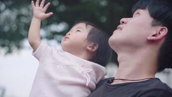 young Asian dad holding his baby girl looking up to the sky waving hand at park under trees, child care parent love bond, children innocence, father and child happy scene, hope for next generation video