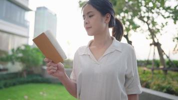 Pretty asian woman reading stories poetry from small book in the relaxing vibe. Fresh green environment and modern high rise building, recreation reading young female, with sunlight leak on background video