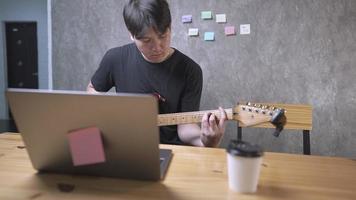 A young guitarist college student practicing on electric guitar reading notes from laptop while doing live streaming, profitable talents, online musical courses at home, using portable laptop computer