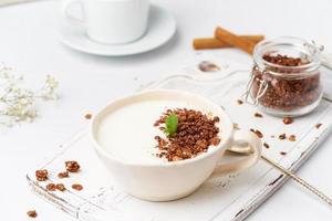 Yogurt with chocolate granola in cup, breakfast with tea on white wooden background, side view.