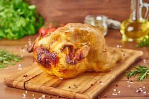 Whole roasted chicken on wooden cutting board on dark brown wooden table, side view photo