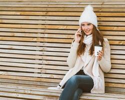 Beautiful young girl drinking coffee, tea from a plastic mug in autumn, winter and talking on a mobile phone. Woman with long hair sitting on a bench in autumn or winter, basking in a hot drink photo