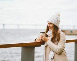 Beautiful young girl drinking coffee, tea from plastic mug in autumn, winter. A woman with long hair stands on waterfront on Baltic sea in port and waiting for ferry, heated by hot drink, copy space photo