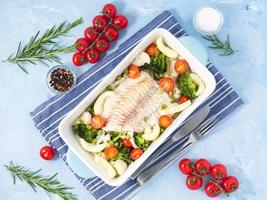 Fish cod baked in blue oven with vegetables - broccoli, tomatoes. Healthy diet food. Blue stone background, top view.