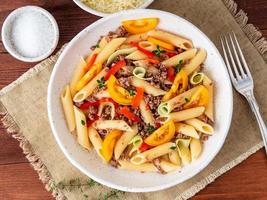 Penne pasta with yellow tomatoes, red and green vegetables, mincemeat on dark wooden background, top view, close up