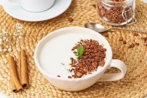 yogur con granola de chocolate en taza, desayuno con té sobre fondo beige, vista lateral, primer plano.
