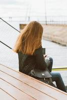 Unrecognizable woman with long hair sits with phone in her hand, her back to camera. Autumn or winter, a girl in jacket outdoors waiting for a call or sms, texting photo