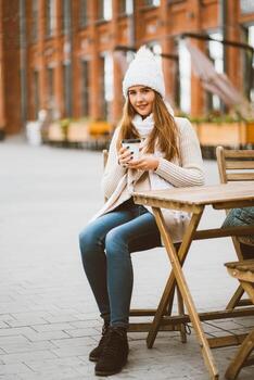 Beautiful young girl drinking coffee, tea from plastic reusable mug in autumn, winter. Woman with long hair, in warm clothes sitting in street cafe, warmed by hot drink, vertical photo