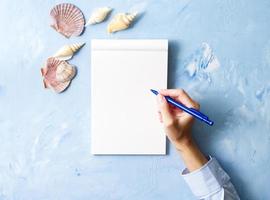 woman writes in notebook on stone blue table, Mock up with frame of seashell, top view photo